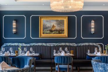 Elegant restaurant interior with navy blue walls, a central nautical painting, plush chairs, and neatly set tables with glassware and napkins. A chandelier hangs from the ceiling.