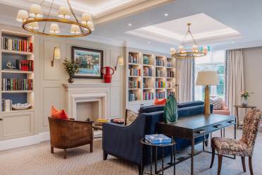Elegantly decorated living room with a blue sofa, armchairs, bookshelves, a fireplace, and ceiling lights. A painting and vase with flowers adorn the mantle. Large windows provide natural light.