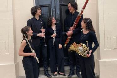 The Dogoda Wind Quintet Five musicians dressed in black stand in front of a door, holding a flute, clarinet, oboe, bassoon, and French horn, and looking at each other.