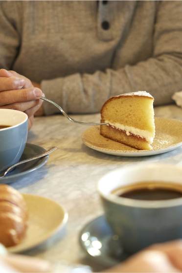 Two people sitting at a table with coffee cups, a slice of cake, and a croissant. A vase with pink and purple flowers is on the table.