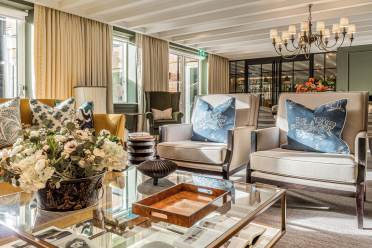 A bright, elegant living room with armchairs, a glass coffee table, floral arrangement, decorative pillows, large windows, and a chandelier overhead.