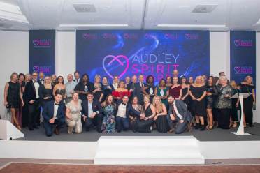 A large group of people in formal attire pose on stage in front of a display that reads "Audley Spirit Awards.