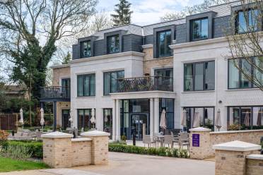 Modern three-story building with large windows and a small balcony on the second floor. Patio umbrellas and seating are visible at the entrance. Trees in the background.