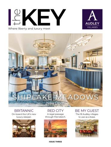 A modern, elegant lounge area with blue chairs, gold accents, and a bar counter, featured on the cover of "The Key" magazine for Audley Villages' Shiplake Meadows opening.