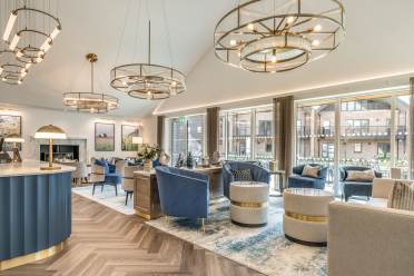 A modern lounge with blue and beige armchairs, round tables, gold light fixtures, large windows, and wood-patterned flooring.