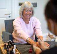 An older woman sits at a table smiling while receiving a manicure from a nail technician in a salon setting.