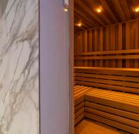 A modern wooden sauna with built-in benches is seen through a glass door with a marble-patterned frame. Warm lighting illuminates the interior.