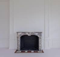 A marble fireplace with a decorative mantel is set against a plain white wall in an empty room with white carpet.