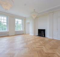 Spacious empty room with herringbone wood floor, large windows, two chandeliers, white walls, and a fireplace.