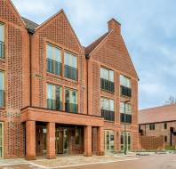 Modern three-story brick apartment building with large windows and glass doors, set against a cloudy sky with another similar building in the background.