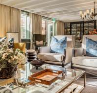A bright, elegant living room with armchairs, a glass coffee table, floral arrangement, decorative pillows, large windows, and a chandelier overhead.