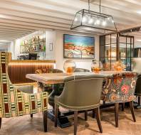 A dining area features patterned armchairs and a wooden table with glass bowls, modern lighting, a bar with bottles, and artwork on the wall.