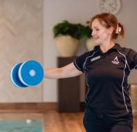 A woman in a uniform holds blue foam dumbbells with arms extended, standing indoors near a pool.