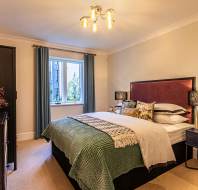 A neatly made bed with green and beige bedding is centered in a bedroom with warm lighting, dark furniture, and a window with blue curtains letting in natural light.
