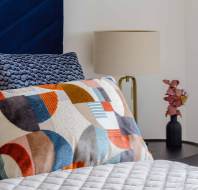 A neatly made bed with colorful geometric pillows, a quilted gray blanket, a bedside table with a lamp, and a small black vase with dried flowers.