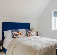 A neatly made bed with a blue upholstered headboard, patterned pillows, beige bedding, and two bedside tables with lamps in a bright, modern bedroom.