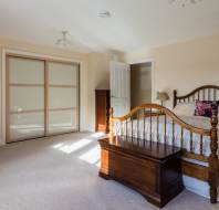 Room with a wooden bed, a chest at the foot, a closed sliding closet, a door slightly ajar, a lamp on a dresser, and sunlight streaming in.