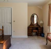 A bedroom with a wooden bed, dresser with oval mirror, armchair, door, and curtained window.