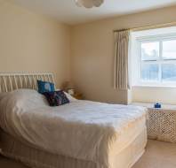 Bedroom with a neatly made double bed, two wooden nightstands, a window with curtains, and a wicker basket below. Walls are beige, and natural light illuminates the room.