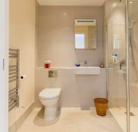 Modern bathroom with beige tiles, featuring a toilet, wall-mounted sink, mirror, towel rack, wicker basket, and a walk-in shower.