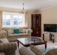 A cozy living room with striped sofas, a glass coffee table, a flat-screen TV, and a corner cabinet. Light streams in through a window with beige curtains. A floral lamp is in the foreground.