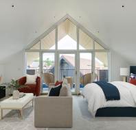 A spacious bedroom with a vaulted ceiling features a bed, a TV, a red couch, a beige loveseat, and a small balcony with outdoor chairs, all under large triangular windows.
