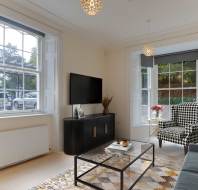 Bright living room with large windows, patterned armchair, black cabinet, wall-mounted TV, glass coffee table, and decorative items. Neutral tones with pops of color in decor.