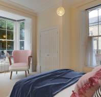 Bright bedroom with a large window, pink chair, and bed with blue and pink bedding. A light fixture hangs from the ceiling, and a building is visible outside through the window.
