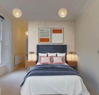 A bedroom with a double bed, blue headboard, and white and blue bedding. Two pendant lights hang from the ceiling. There are two framed artworks above the bed and two nightstands with lamps.