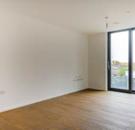 Empty room with wooden floors, white walls, and large floor-to-ceiling window. Urban view outside.