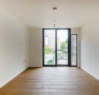 An empty room with wooden flooring, white walls, and a large window leading to a balcony with a view of buildings and trees outside.