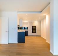 A modern kitchen with light wood floors, white walls, and recessed lighting. Features include a built-in oven, a refrigerator, and a minimalist design.