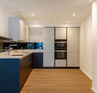 Modern kitchen with wooden floors, white walls, and recessed lighting. Features a sleek black counter, built-in double oven, and minimalist cabinets.