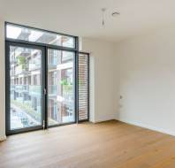 Empty room with large floor-to-ceiling windows, wooden floors, and a view of a modern apartment building exterior.