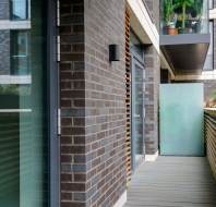 A modern building exterior with brick walls, glass doors, and a wooden deck. A small potted plant is visible on a balcony above.