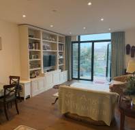 A cozy living room with a dining table, chairs, built-in shelves, a TV, and a couch facing sliding glass doors that lead to a patio outside. The space is decorated with paintings and plants.