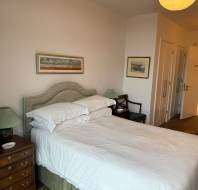 A neatly made bed with white linens is in a bedroom. There are two nightstands with lamps, a chair, and framed artwork on the walls. A partially open door reveals a bathroom.