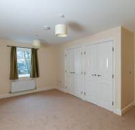 Empty room with beige carpet, cream walls, and two pendant lights. A window with green curtains on the left. Two sets of white double doors on the right wall.