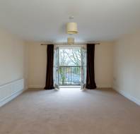 An empty room with beige walls and carpet, featuring a ceiling light and double doors leading to a small balcony with brown curtains. A radiator is mounted on the left wall.