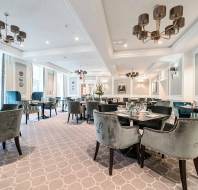Elegant, modern restaurant dining room with velvet chairs, set tables, neutral décor, large windows, chandeliers, and artwork on the walls.