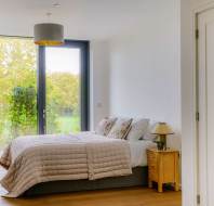 A bedroom with a double bed, a bedside table with a lamp, and large windows showing green foliage. White walls and wooden flooring create a minimalistic design.