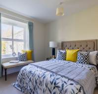 A bedroom with a bed featuring a patterned duvet and colorful pillows. Two bedside tables with lamps are on either side. A window with blue curtains lets in natural light.