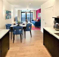 Modern open-plan kitchen and living area with wood flooring, black and white cabinetry, a dining table with blue chairs, and a blue sofa. Colorful artwork and decor accent the space.