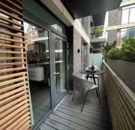 Narrow balcony with wooden flooring and slatted walls, featuring a small table, two chairs, and potted plants, adjacent to a glass door leading inside a building.