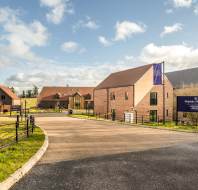 A modern residential development with brick houses on a sunny day. Flags with development name line the road. Blue sky and clouds are visible.