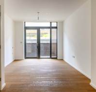 Empty room with wooden floor, white walls, and a ceiling light. Large glass doors lead to a small balcony with slatted wooden panels, allowing natural light to fill the space.