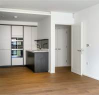 Modern kitchen with built-in appliances, sleek cabinets, and a small peninsula. The kitchen is adjacent to an open area with wooden flooring and a partially open white door.
