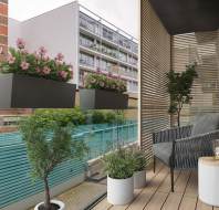 Modern balcony with two planters of pink flowers, a gray cushioned chair, a small round table with a candle, and a potted plant. Wood slat wall and glass railing overlook neighboring buildings.