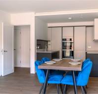 Modern kitchen with stainless steel appliances, dark cabinets, and a wooden dining table surrounded by blue chairs. White walls and wood flooring complete the space.