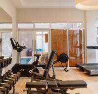 A gym with various equipment including dumbbells, exercise bikes, and treadmills. A mirrored wall and a view of a pool area through glass doors in the background.